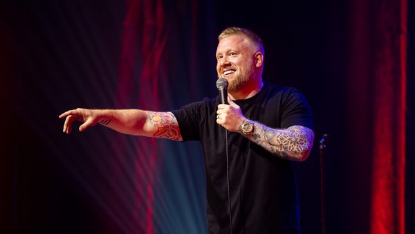 A man wearing a black t-shirt holding a microphone on stage, he is smiling and looking out towards with audience with his hand pointing out in front of him.