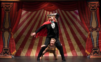 Two performers on stage, in front of a backdrop of a circus tent with red velvet curtains.