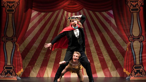 Two performers on stage, in front of a backdrop of a circus tent with red velvet curtains.