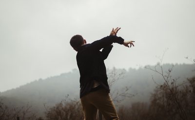 A photograph of a dancer standing with their back to the camera facing out towards a misty tree covered hill. The dancer is wearing dark trousers and a dark top and has their arms stretched out to their right in mid movement.