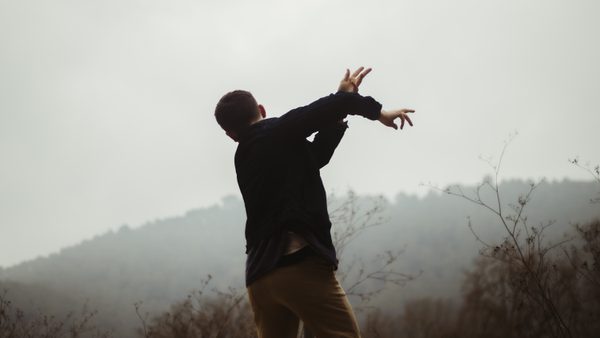 A photograph of a dancer standing with their back to the camera facing out towards a misty tree covered hill. The dancer is wearing dark trousers and a dark top and has their arms stretched out to their right in mid movement.
