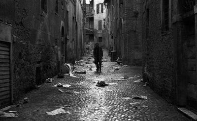 A black and white photo of an alleyway littered with paper and rubbish, a man walks towards the camera.