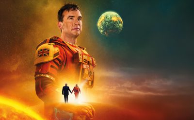 A person in red‑orange futuristic armour stands in a glowing sci‑fi landscape, with a large planet overhead and two smaller figures walking hand‑in‑hand toward the bright horizon.