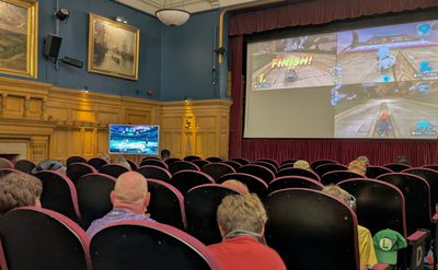 A photo of rows of seats facing towards a large screen, people are sat in the chairs watching as a Nintendo game is projected on to the screen.