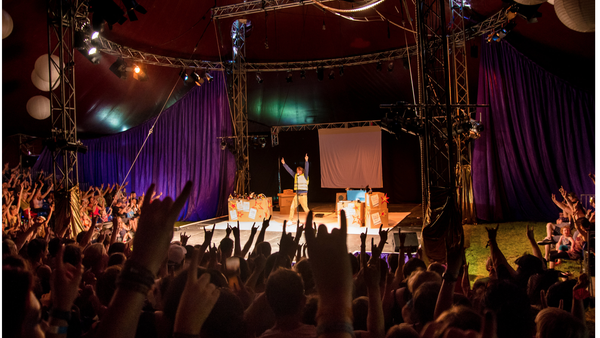 A performer stands on a brightly lit stage inside a large tent, arms raised toward a packed crowd who are also lifting their hands. The venue has a red ceiling, purple curtains and metal trusses with stage lighting. Colourful props with stars and signs sit around the stage, and the atmosphere is energetic and celebratory.