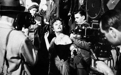 A woman in an elegant gown performs on a classic film set, surrounded by crew and vintage equipment.
