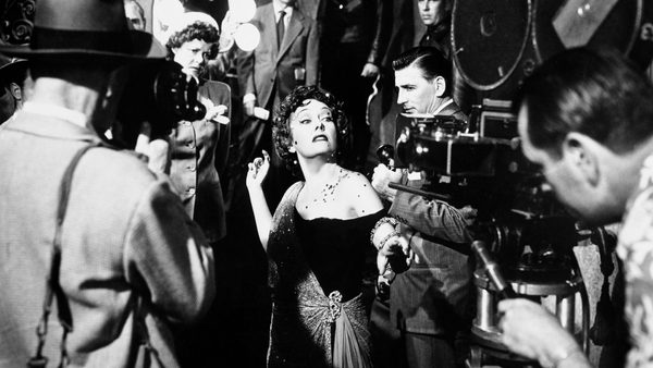 A woman in an elegant gown performs on a classic film set, surrounded by crew and vintage equipment.