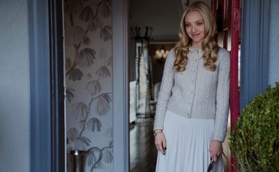 A person with long blond hair stands in a doorway, wearing a light cardigan and skirt. They hold a small knife and wear a crystal-like crown. A red door and leafy wallpaper are visible.