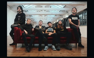 Group of six people dressed in black clothing, sitting and standing around a red sofa in a spacious room with wooden floors and white beams.